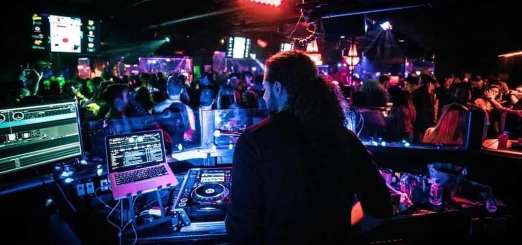 DJ with curly hair operating equipment in a dimly lit club.