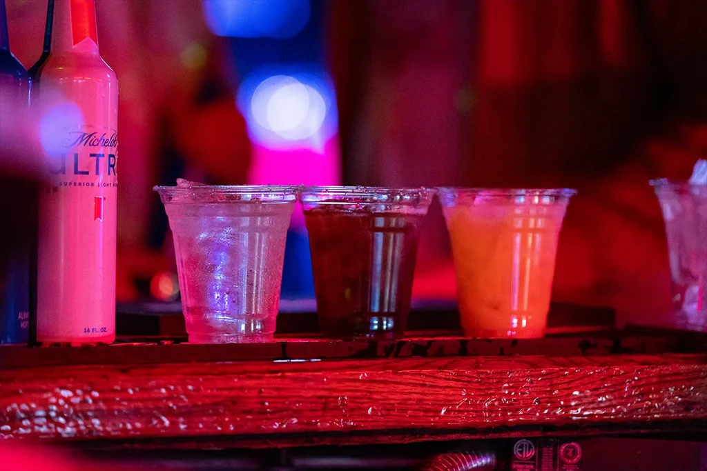 Michelob Ultra bottle and three colorful drinks on a bar.