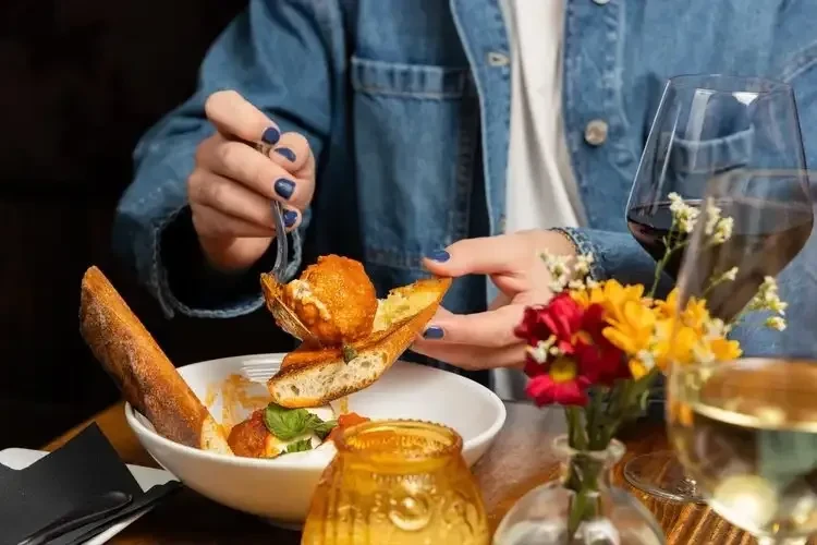 Person's hands with food, wine, and flowers on a restaurant table.