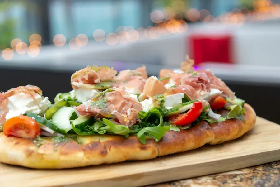 Gourmet flatbread with prosciutto, burrata, arugula, cucumber, and tomatoes.