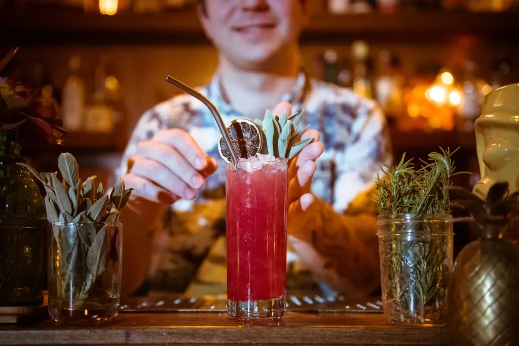 Bartender garnishes a red cocktail with an orange slice and herbs.