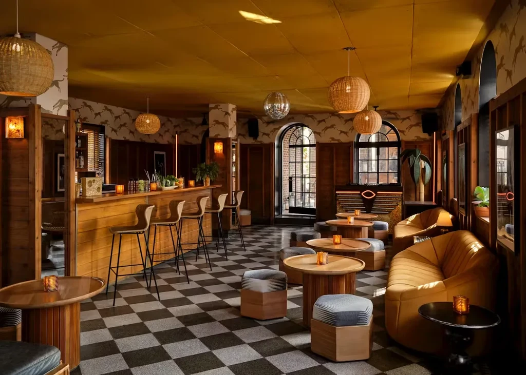 Warmly lit bar with wooden counter, patterned wallpaper, and checkered floor.