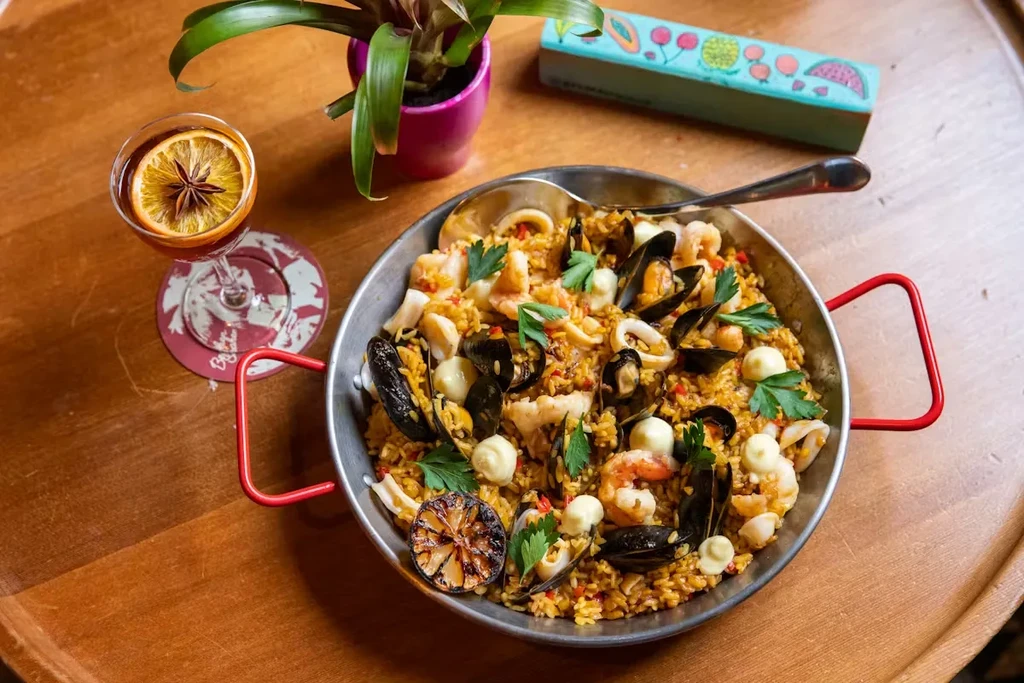 Seafood paella in a pan with a drink on a wooden table.