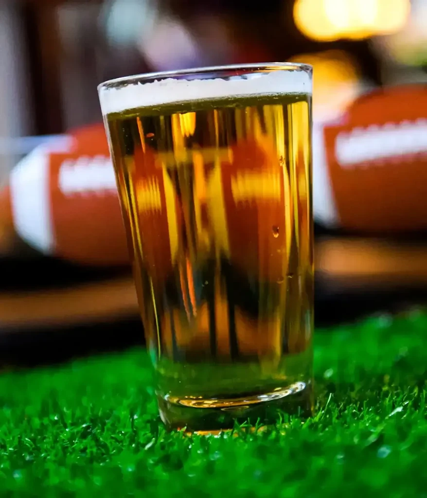 Glass of amber beer on artificial grass, blurred footballs background.