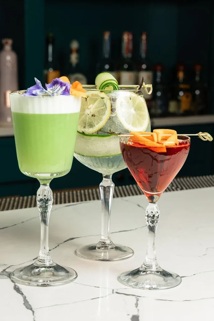 Three garnished cocktails, one green, one clear, and one red, on marble.