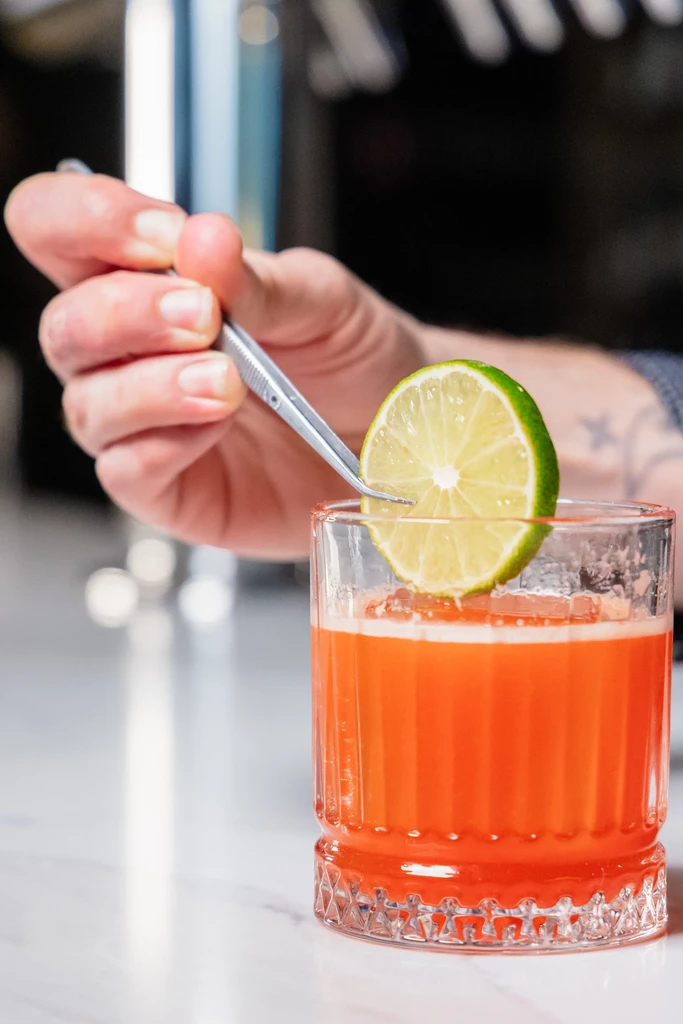 Hand garnishes an orange cocktail with lime using silver tweezers.
