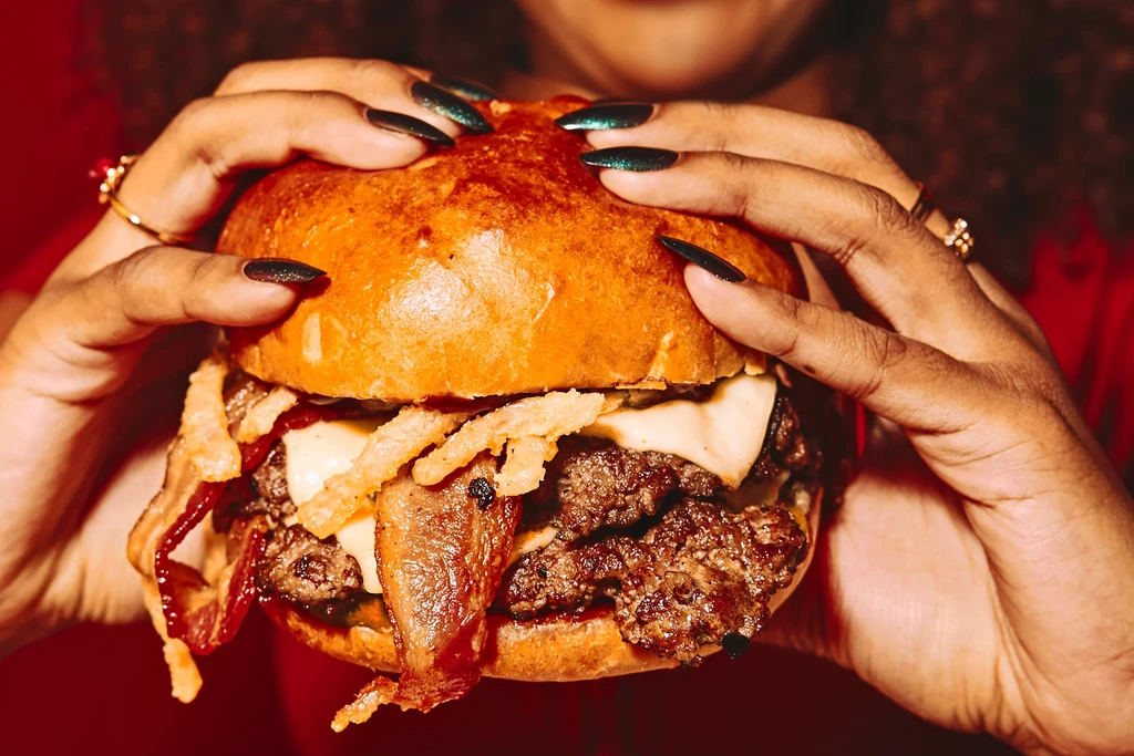 Person holding a large burger with bacon, cheese, and onion strings.