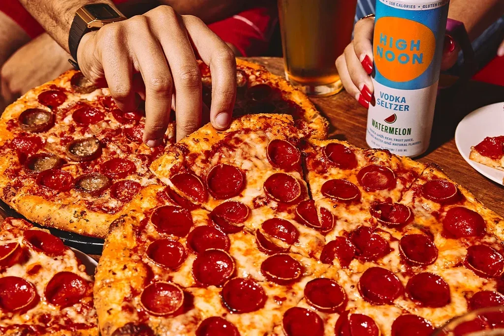 Hands grabbing pepperoni pizza slices, holding a watermelon hard seltzer.