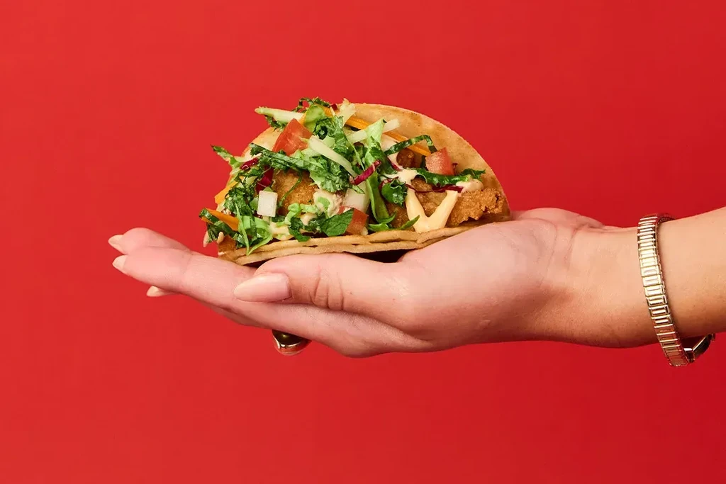 Hand holds a soft-shell taco filled with crispy items and salad.