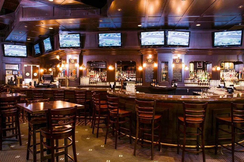 Dimly lit bar with long counter, stools, and wall-mounted TVs.