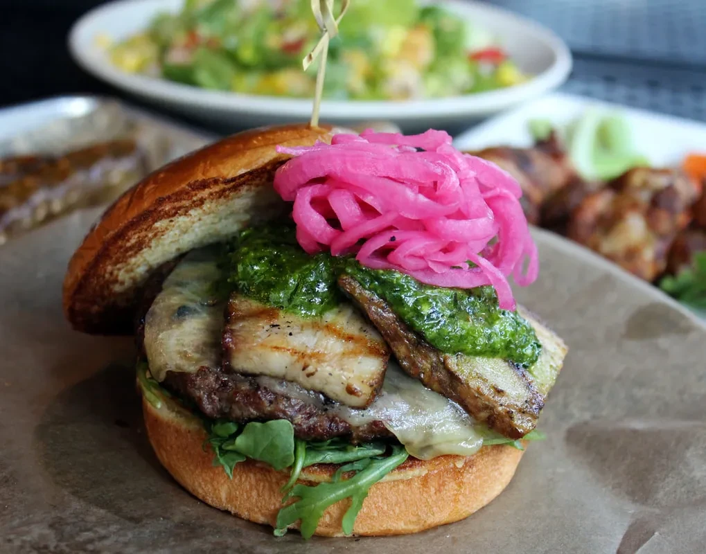 Gourmet burger with patty, melted cheese, pork belly, pesto, pickled onions.