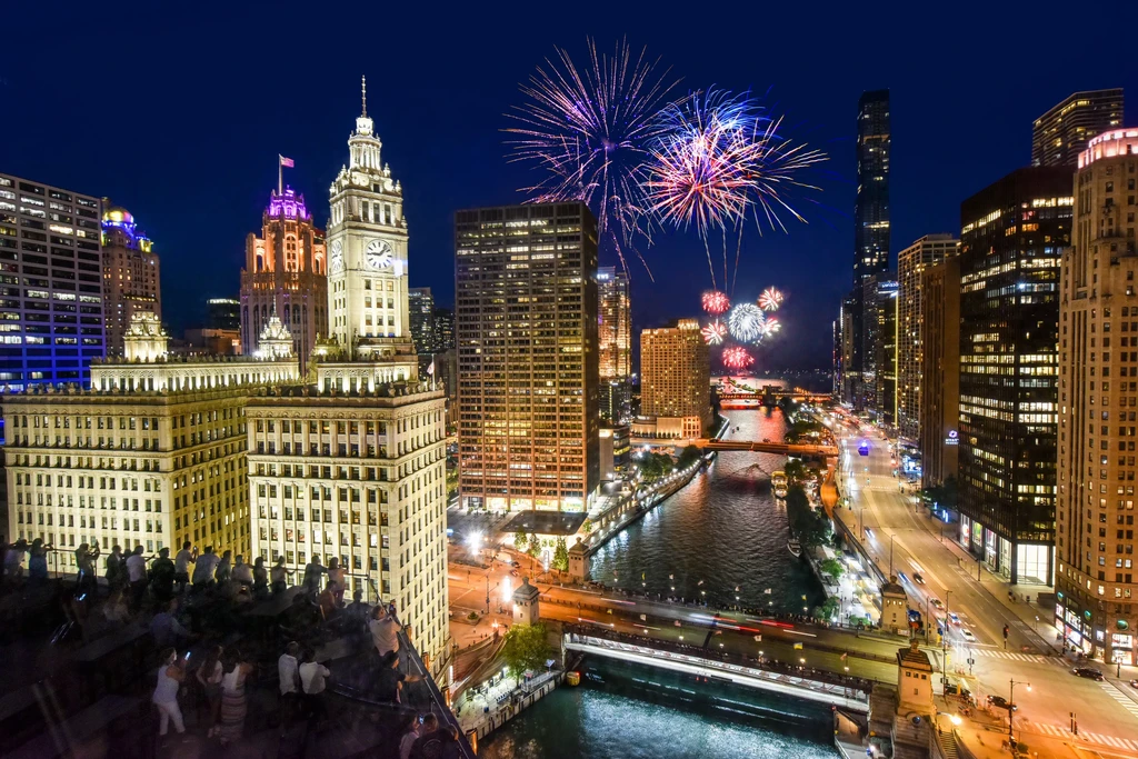 Night cityscape with fireworks over a river, viewed by a crowd.