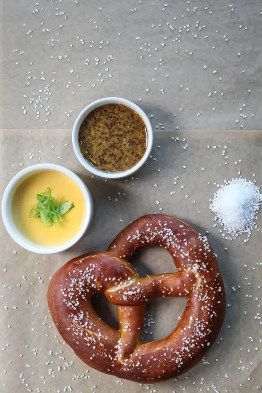Overhead view of a pretzel with mustard, cheese sauce, and salt.