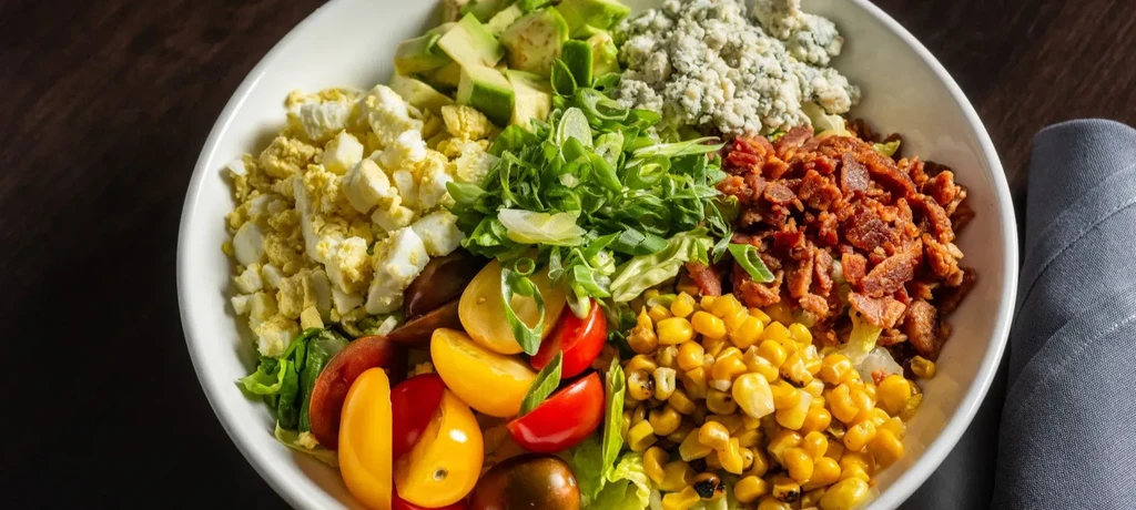 A white bowl filled with sections of hard-boiled eggs, avocado, blue cheese, bacon, corn, tomatoes, and scallions.