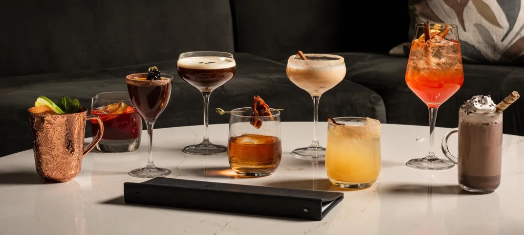 Eight assorted garnished drinks in various glassware on a reflective surface.