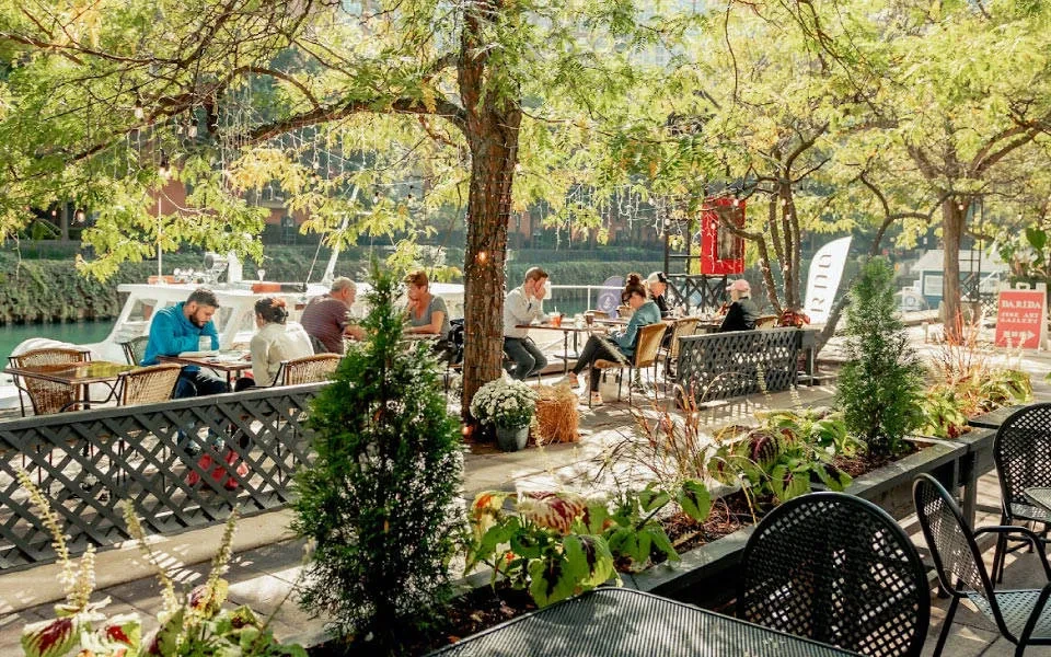Outdoor restaurant patio with patrons under dappled sunlight near water.