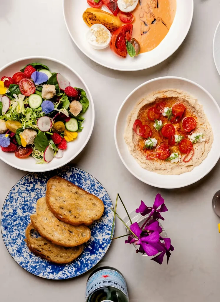 Flat lay of salads, hummus, tomatoes, bread, and a drink.