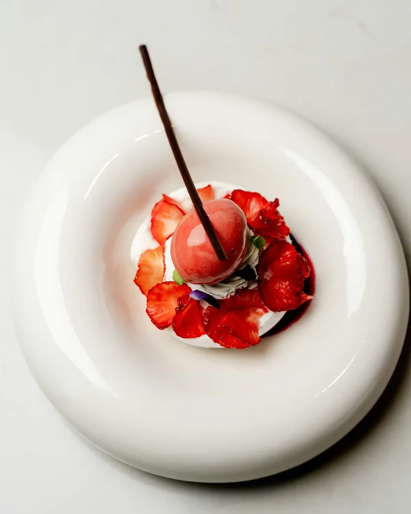 Dessert in a white bowl, topped with strawberries, sorbet, and chocolate.