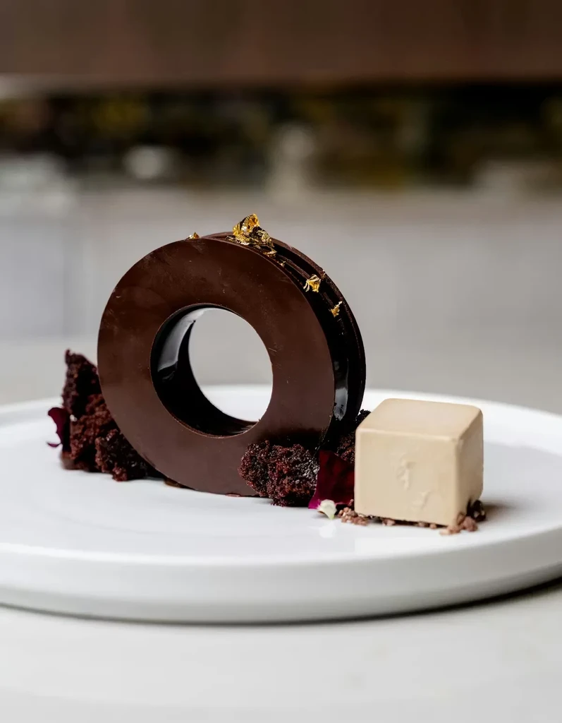 Artistic chocolate dessert with gold leaf, cake, and beige cube.
