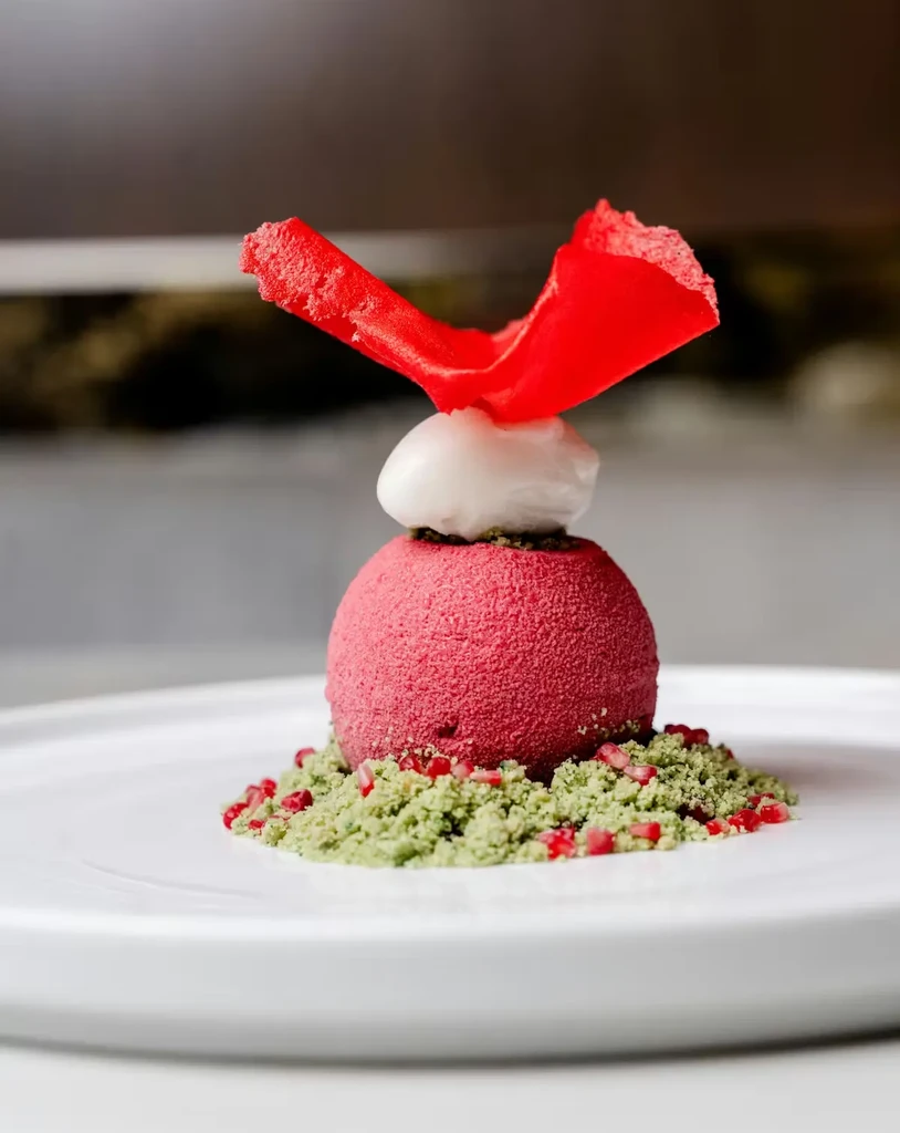 Magenta spherical dessert with white cream, red garnish, and green crumble.