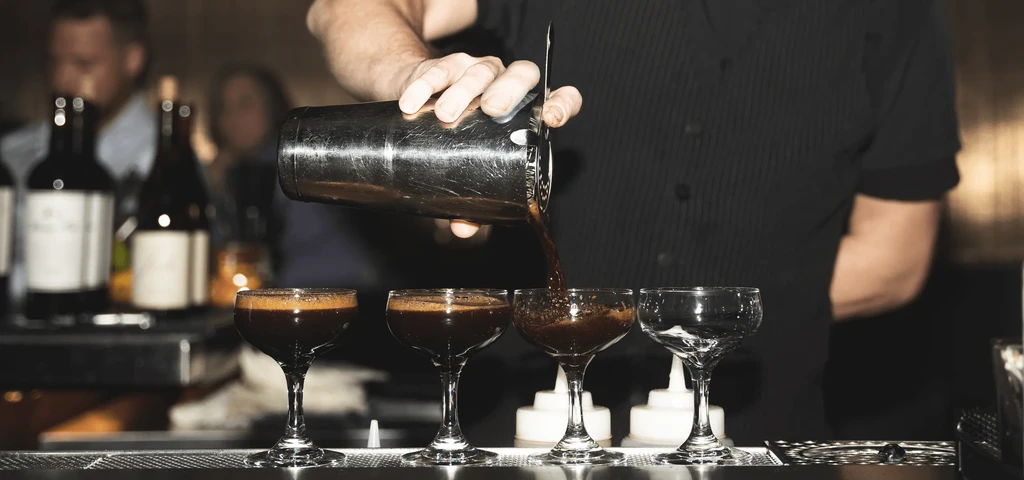 Bartender pours dark beverage from shaker into stemmed glasses.