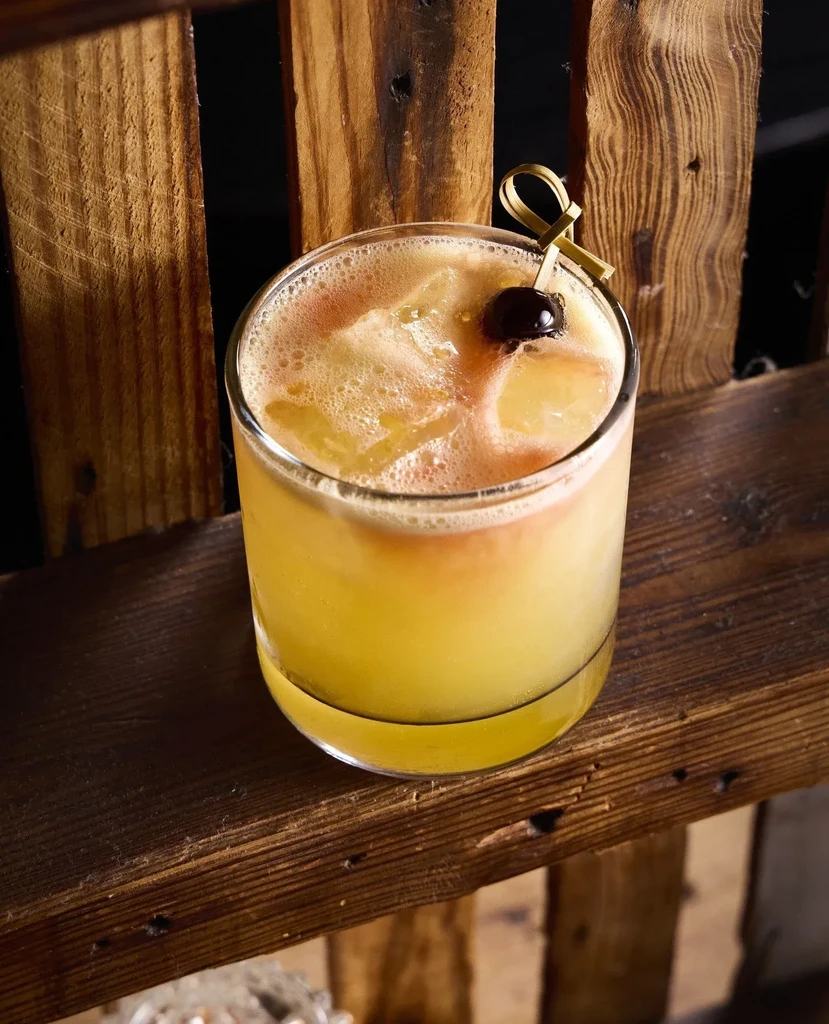 Yellow cocktail in short glass, dark cherry, wooden surface.