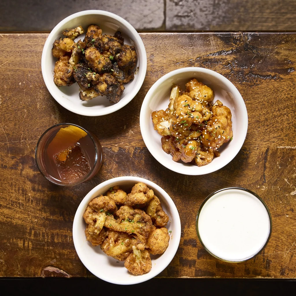 Three bowls of cauliflower dishes, two drinks on a wooden table.