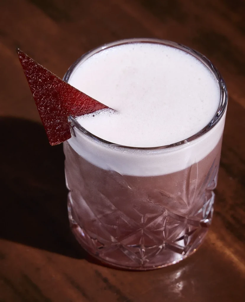 Cocktail in a diamond-patterned glass with foam and red garnish.