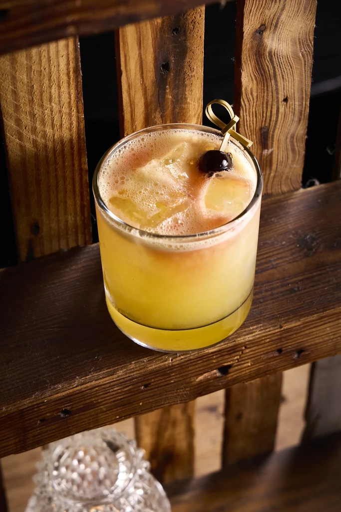 Vibrant cocktail with foamy top, ice, cherry on rustic wooden surface.