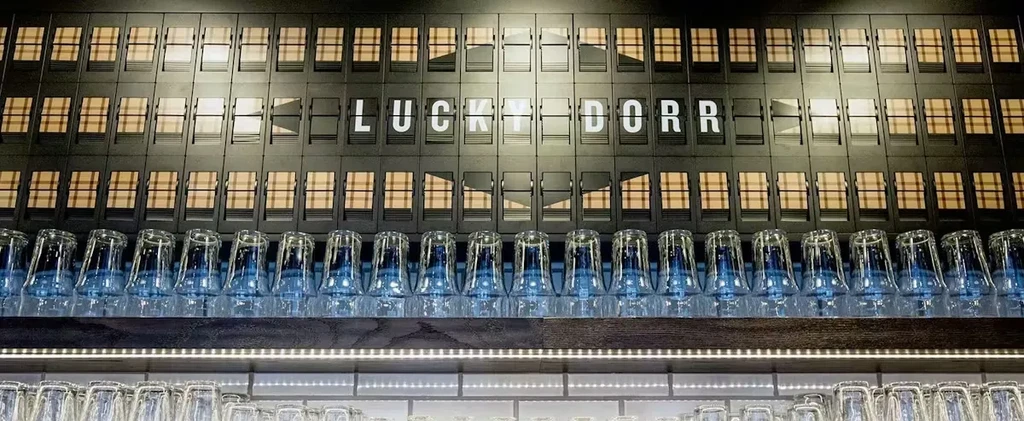 Split-flap display shows "LUCKY DORR" above shelves of inverted glasses.