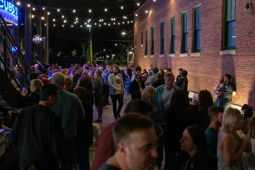 Evening crowd between modern and brick buildings with string lights.