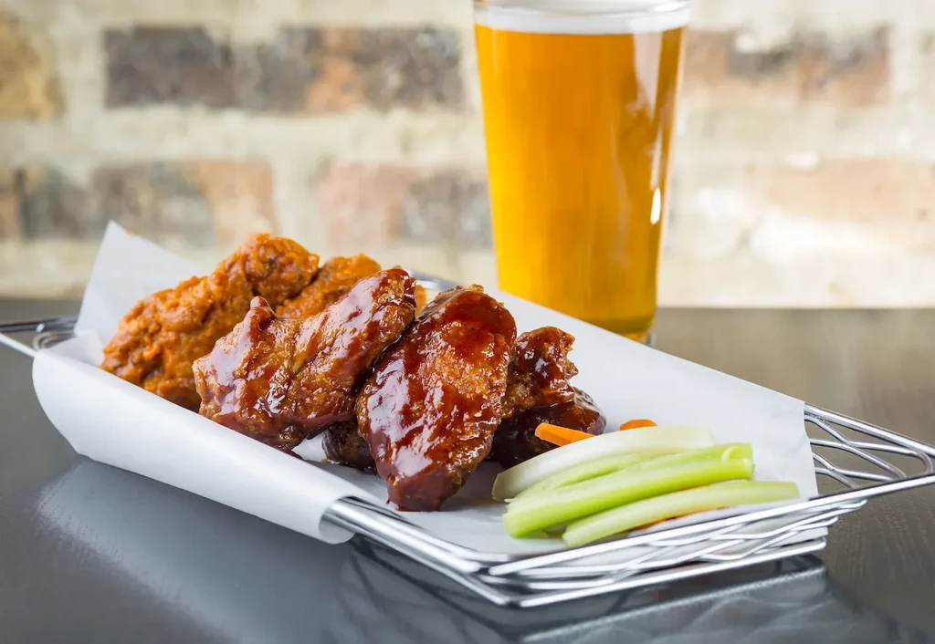 Basket of buffalo and BBQ chicken wings with celery and beer.