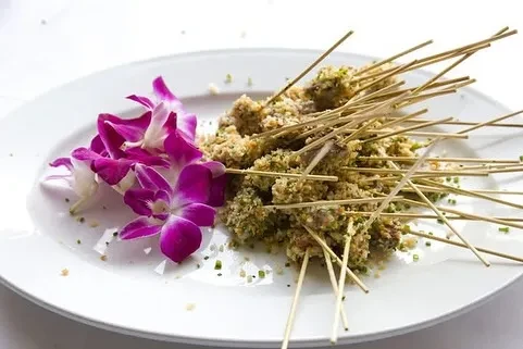 Skewers with green coating, purple orchids, and chives on a white plate.