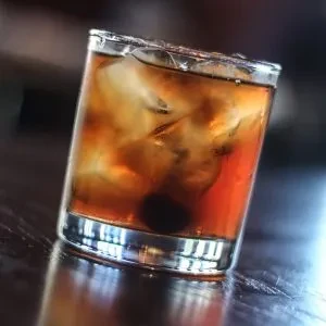 Glass of dark amber cocktail with ice and a cherry.