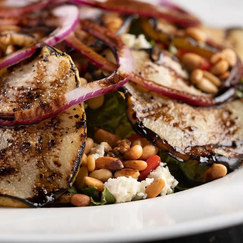 Close-up of a salad with grilled vegetables, red onion, pine nuts, and cheese.