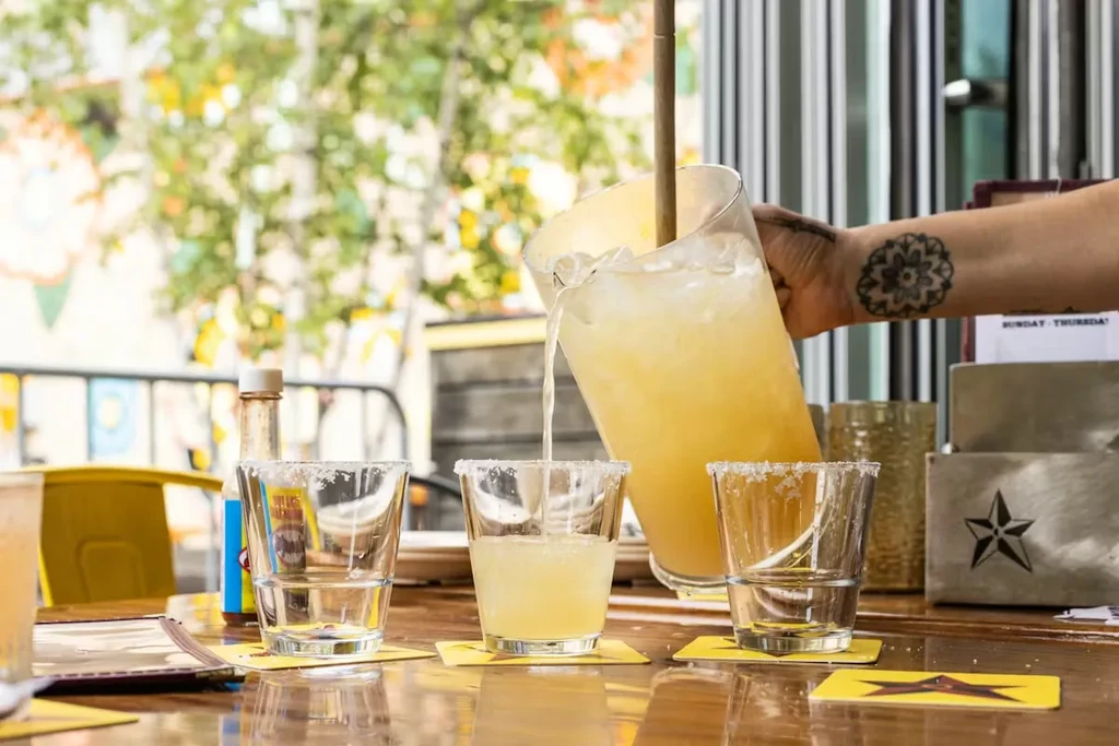 Hand pours yellow iced beverage from pitcher into salt-rimmed glass.