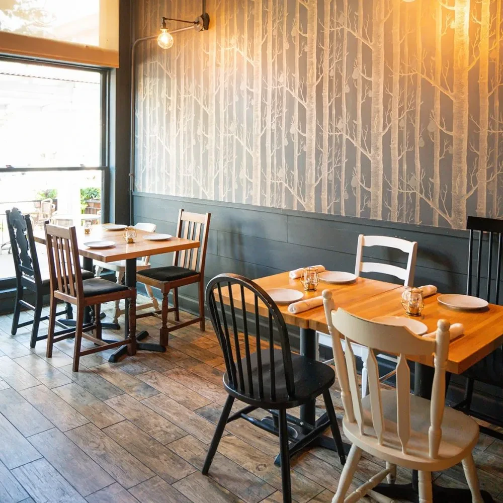 Restaurant dining area with light wood tables and tree-patterned wallpaper.