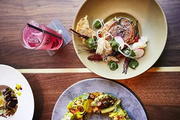 Overhead view of three dishes and a pink cocktail on a wooden table.