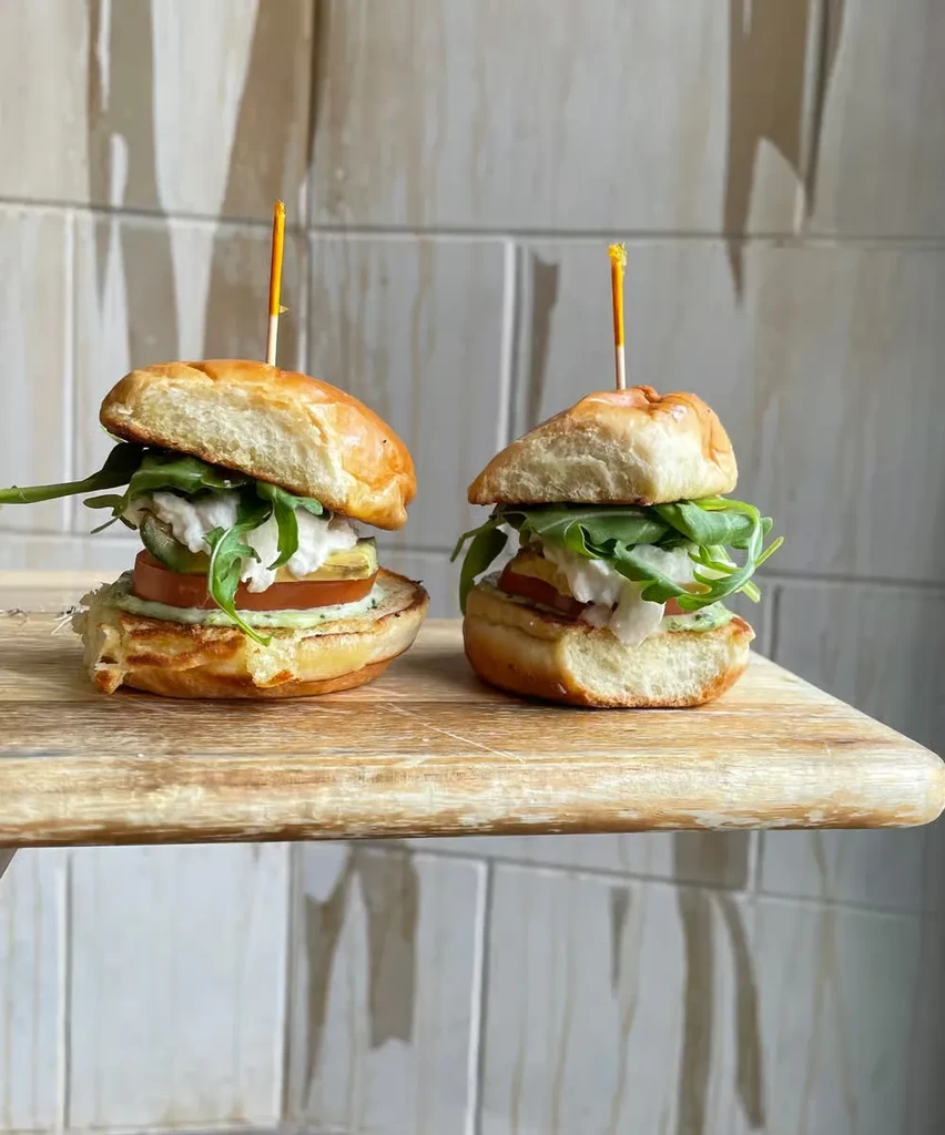 Two gourmet sliders with arugula, tomato, and creamy sauce on a board.