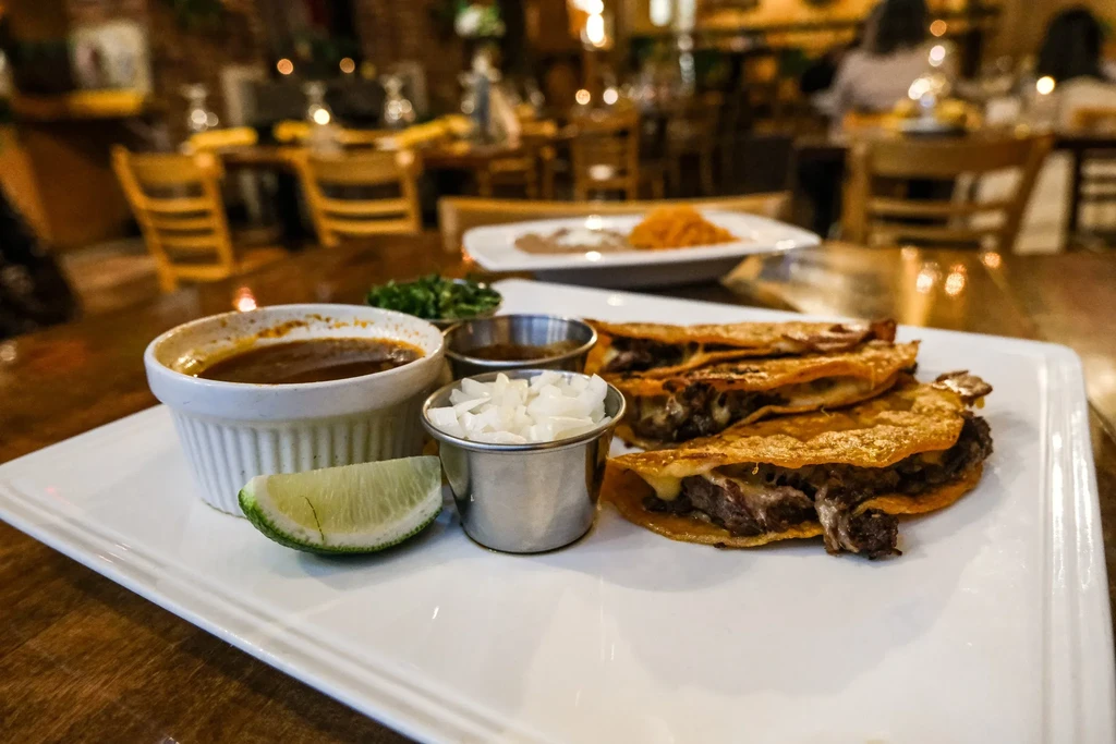 Three birria tacos with consommé, lime, and onions on a plate.