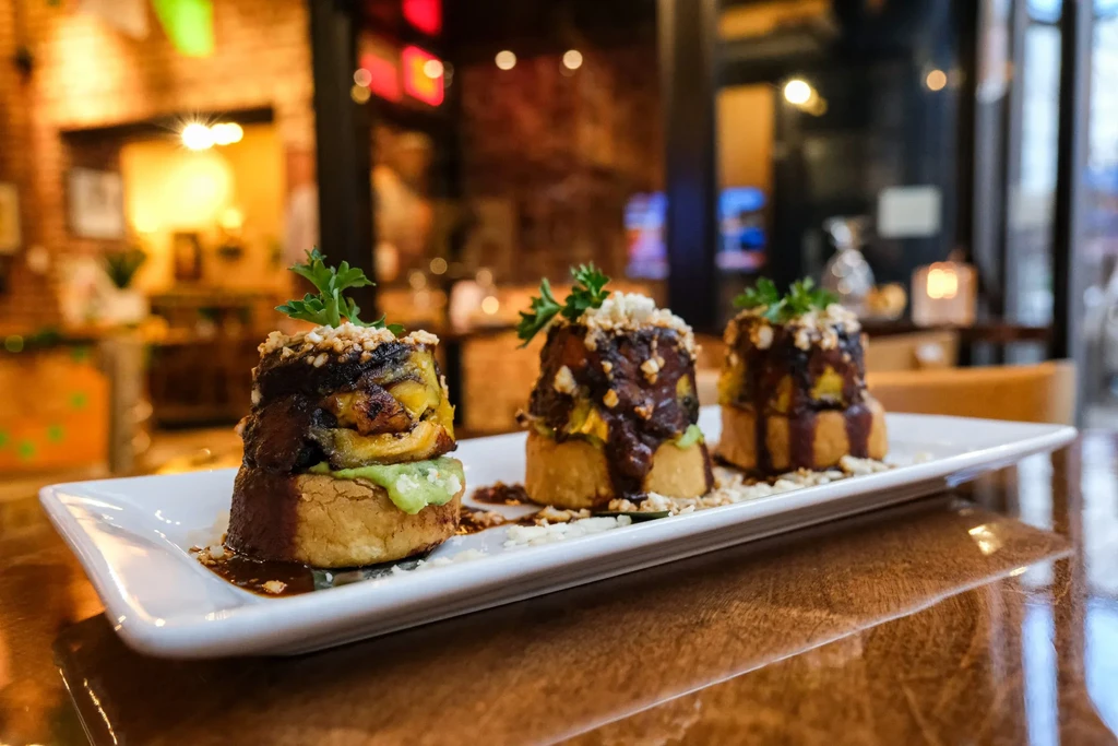 Three appetizers with plantain, green sauce, and nuts on a white plate.