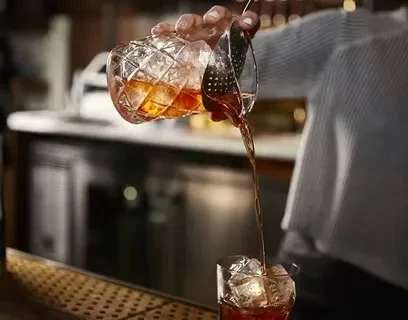 Bartender pours amber liquid from mixing glass into ice-filled glass.