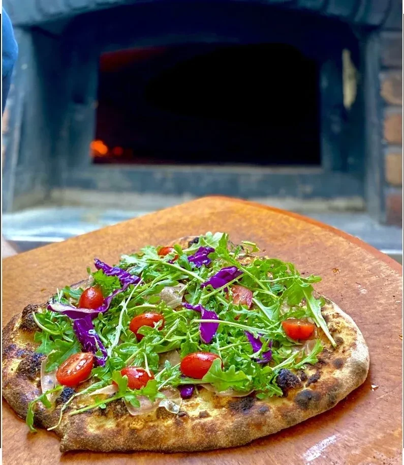 Wood-fired pizza with arugula, tomatoes, and prosciutto on a wooden peel.