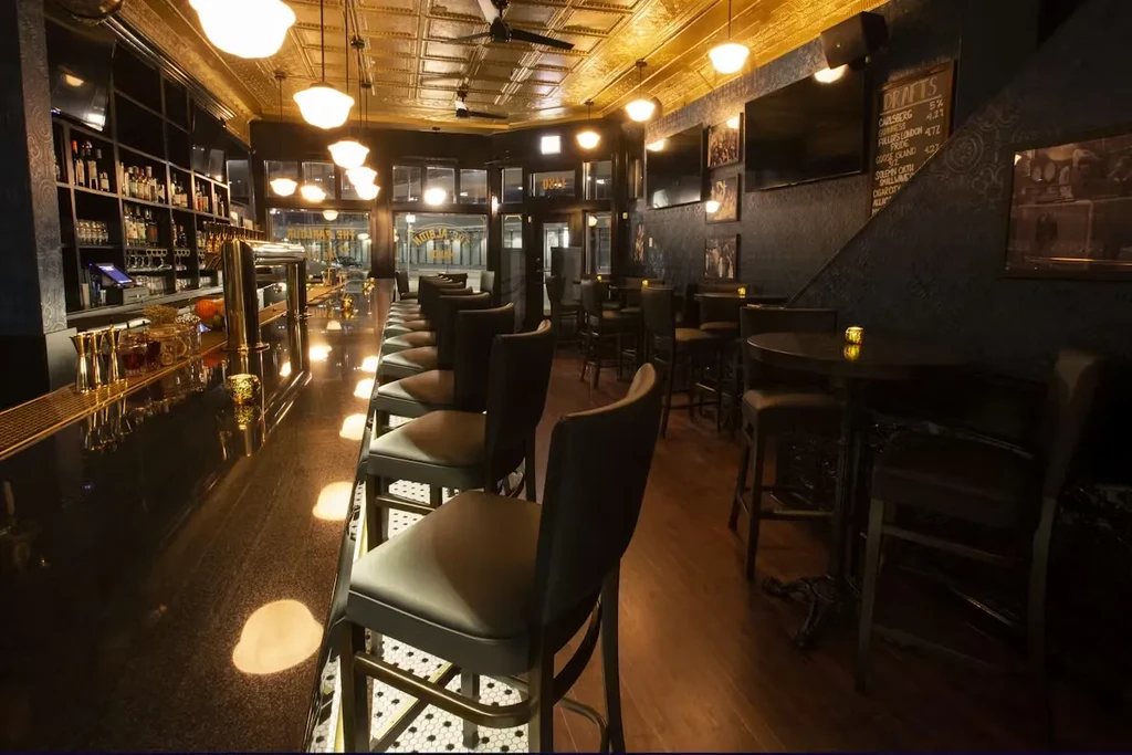 Dimly lit upscale bar interior with long counter, bottles, and tables.