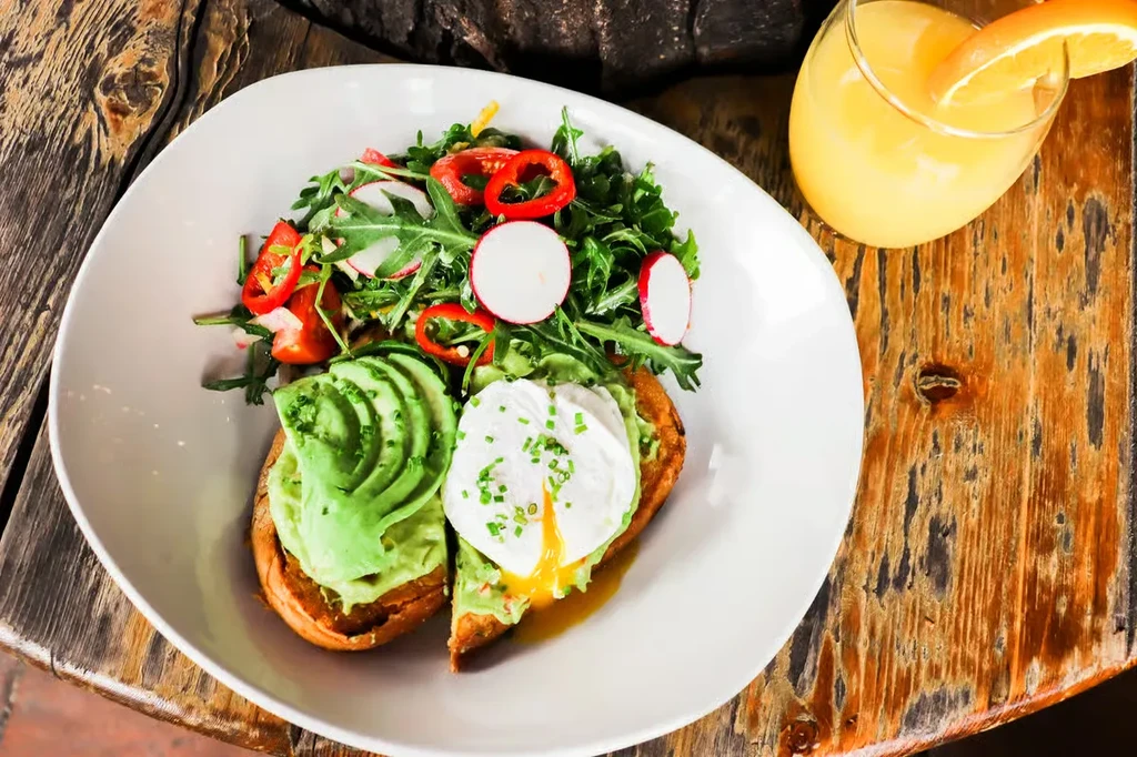 Gourmet breakfast with avocado toast, poached egg, salad, and orange juice.
