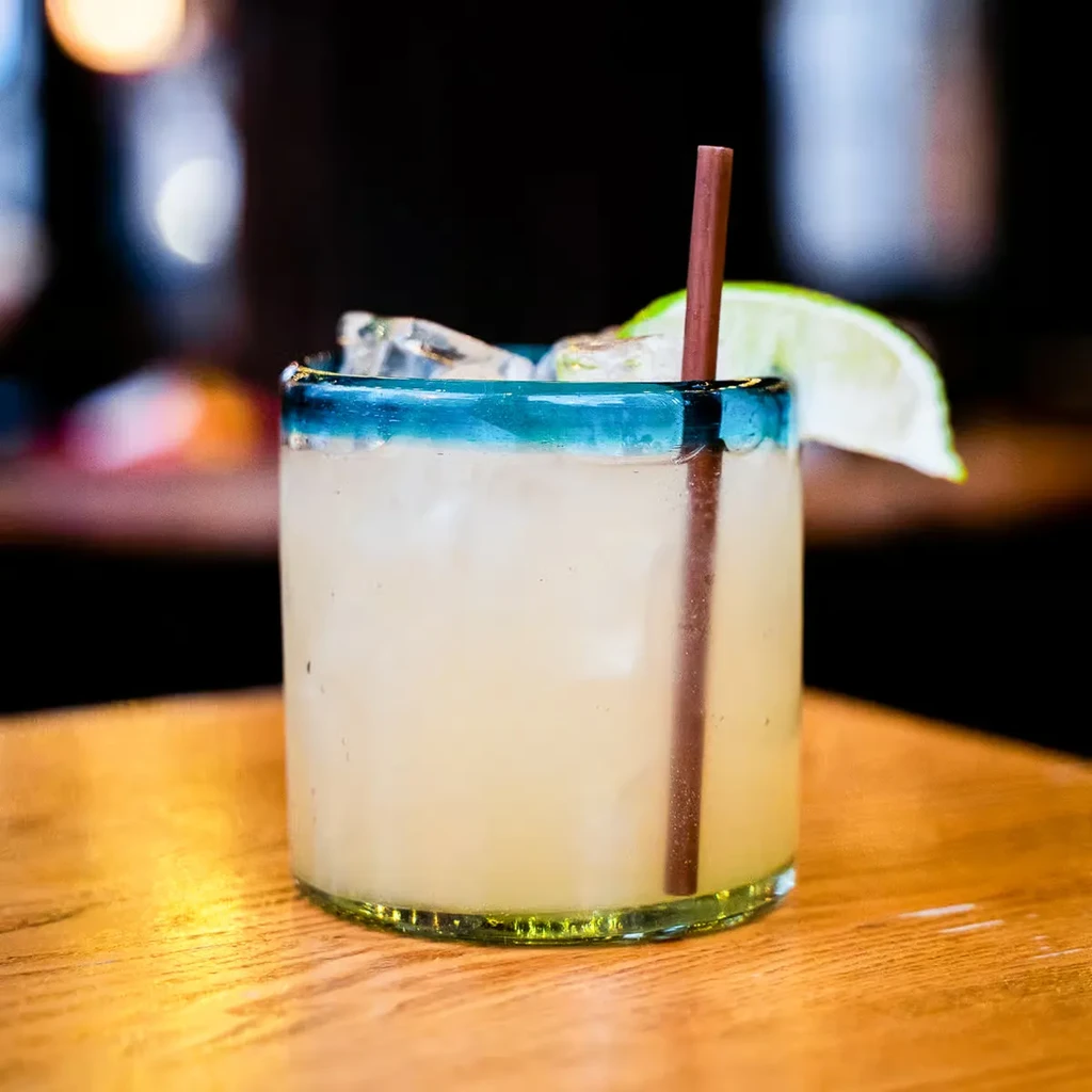 Pale yellow cocktail with ice, lime, and teal blue rim.