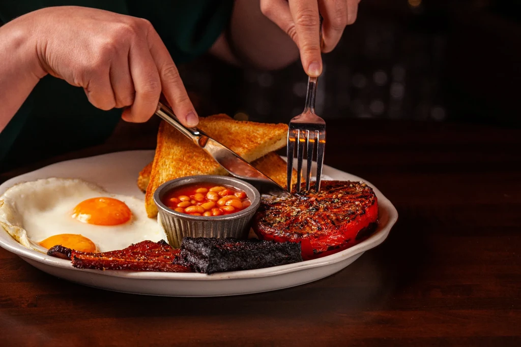 Hands cut into a Full English breakfast with knife and fork.