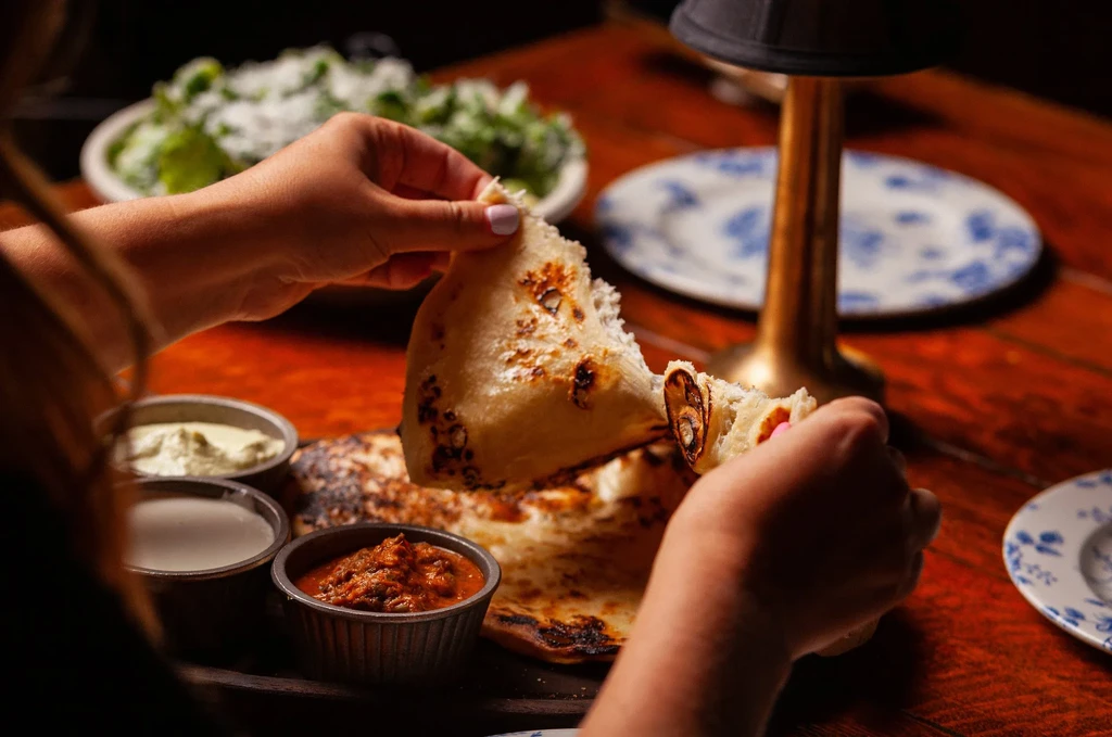 Hands tearing naan bread, surrounded by dips and salad.