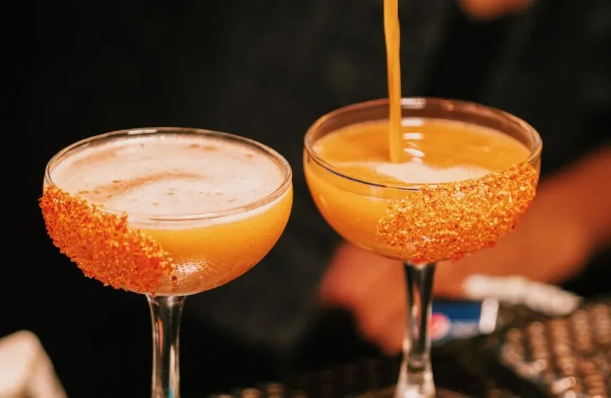 Two orange cocktails with sugar rims are being poured and served.