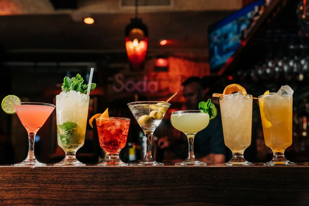 Seven vibrant cocktails are arranged on a dark wooden bar counter.
